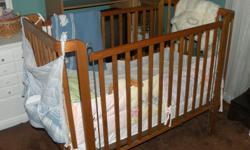 Oak crib with mattress. Accessories included are bumper pad, sheets, blankets, cloth diaper container and baby product container. Sides of crib can be lowered.